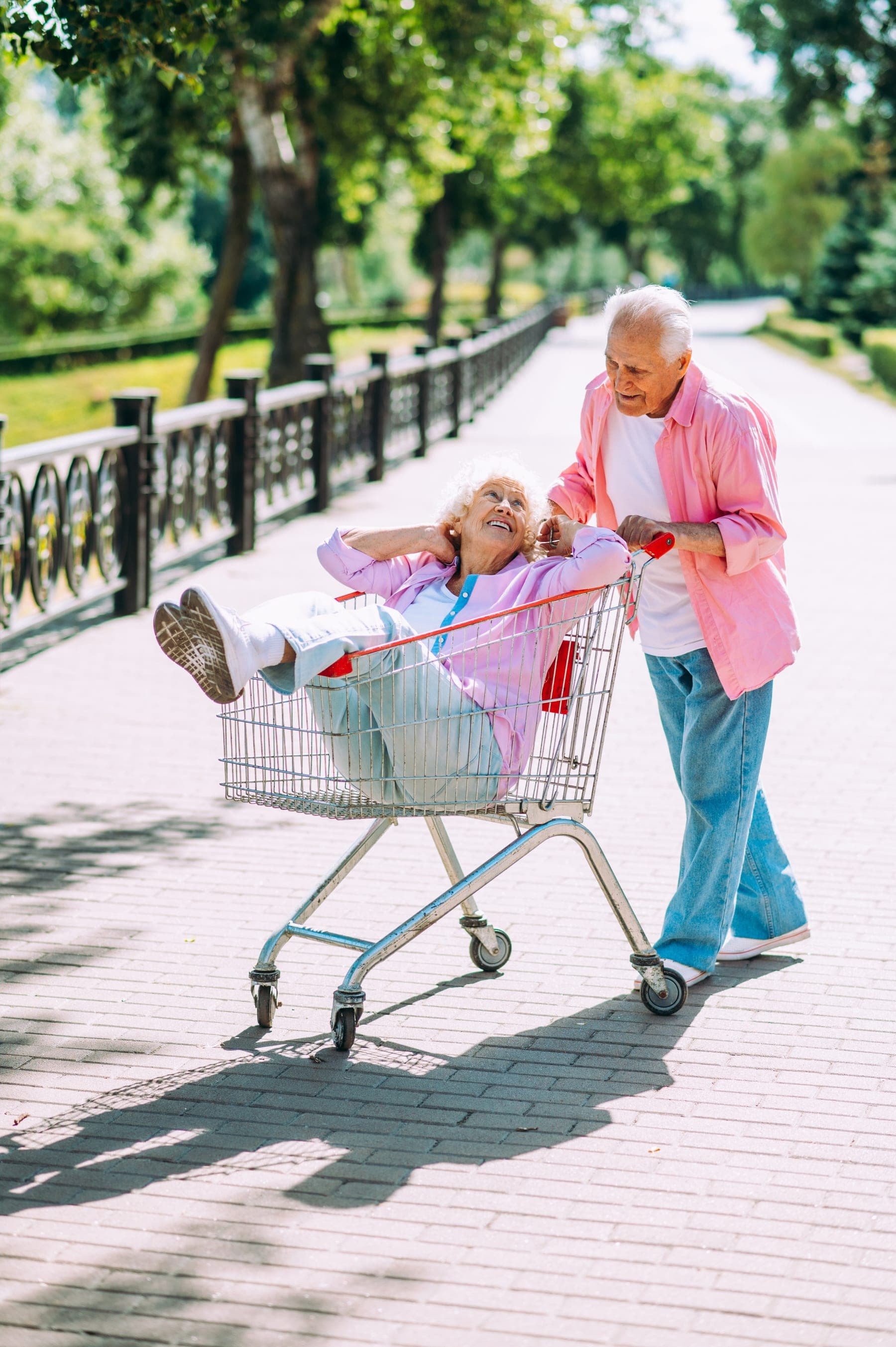 Eine Seniorin sitzt lachend im Einkaufswagen, ihr Mann schiebt sie durch den Park