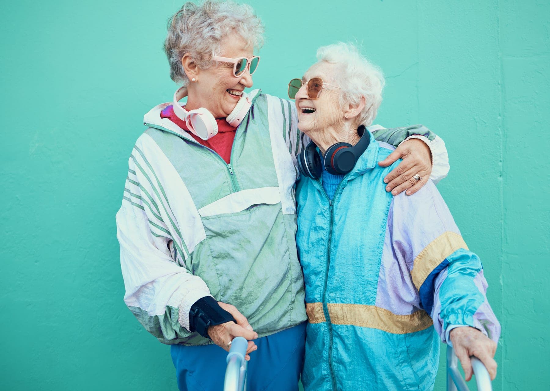 Zwei Seniorinnen in bunten Trainingsjacken lachen sich an, eine mit Gehhilfe